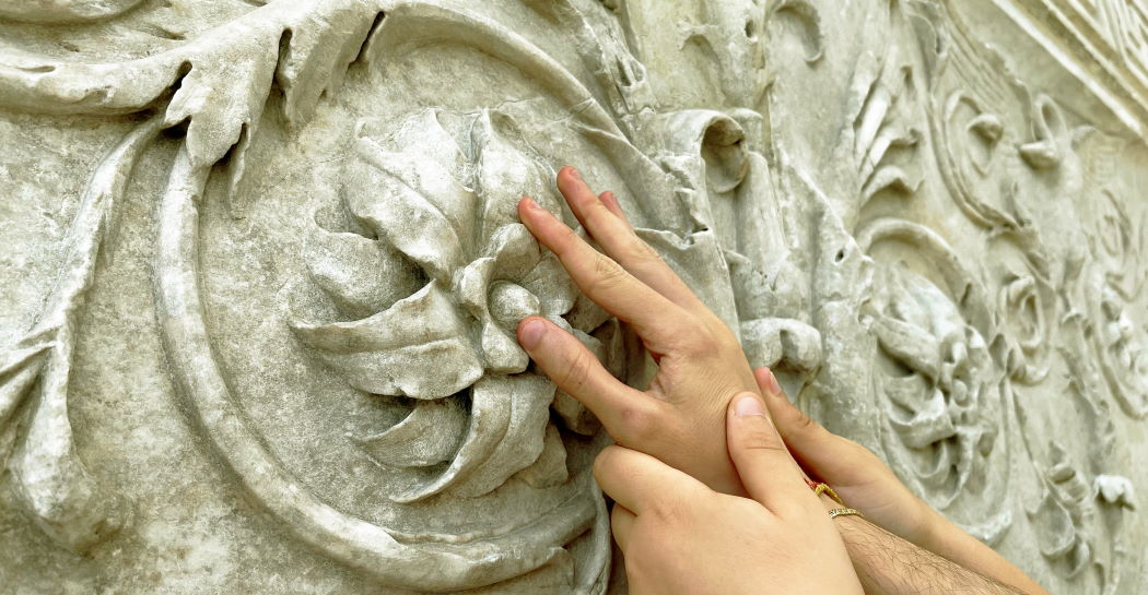 Mani che toccano una decorazione vegetale dell’Ara Pacis